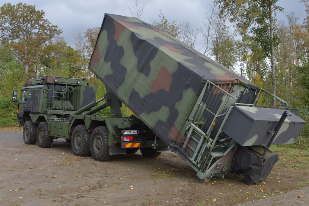 Die beauftragten Standard-Wechselpritschen werden bereits in der Bundeswehr genutzt, hier wird die Wechselpritsche als Ladungsträger für einen Container genutzt und gerade von einem Lkw, der als Trägerfahrzeug dient, abgesetzt. Blauer Bund